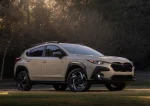 Передній ракурс 3/4 Subaru Crosstrek Hybrid 2026 року пісочного кольору на фоні вечірнього лісу.
