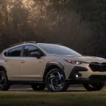 Передній ракурс 3/4 Subaru Crosstrek Hybrid 2026 року пісочного кольору на фоні вечірнього лісу.